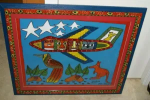 RARE UNIQUE COLORFUL  FOLK ART PAINTING PAPUA NEW GUINEA HUMOROUS ARTIST: TRIBAL WARRIORS TRAVELLING BY AIRPLANE & FRAMED IN SIGNED HAND PAINTED FRAME TO MATCH THE ART DESIGNER COLLECTOR WALL CARTOON  ART  26 1/2” by 23 1/2” HUGE DFP8